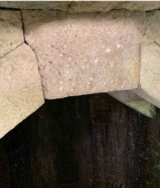 Close-up of a stone arch made from light-coloured rectangular blocks, set above a dark, shadowy space. The stones appear weathered with visible surface texture and mortar lines between them.