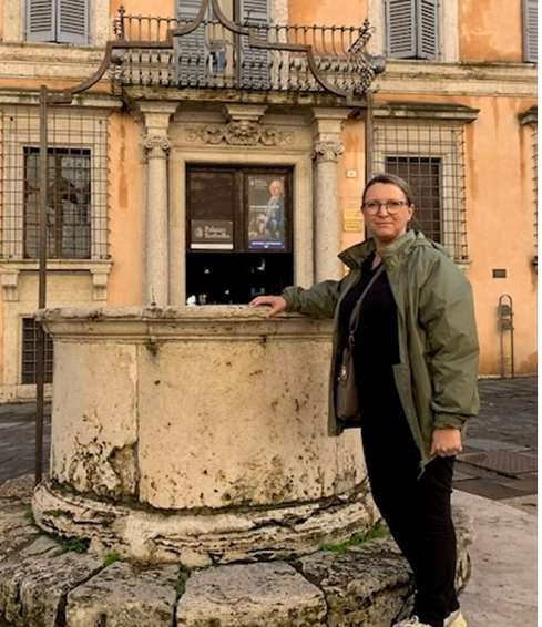 A person in a green jacket stands beside an old stone well in a European square, with a historic building featuring ornate windows and a poster in the background.