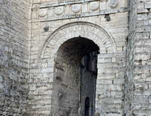 Stone archway with Latin inscription AVGVSTAN PERUSIA, featuring circular carvings above and surrounded by weathered stone walls; a building is visible through the opening, showcasing enduring craftsmanship reminiscent of classic fabrication.