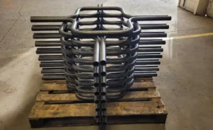 Stacked metal pipes with curved ends, produced using CNC Tube Bending, are arranged neatly on a wooden pallet in a warehouse, with an open garage door and part of a car visible outside.
