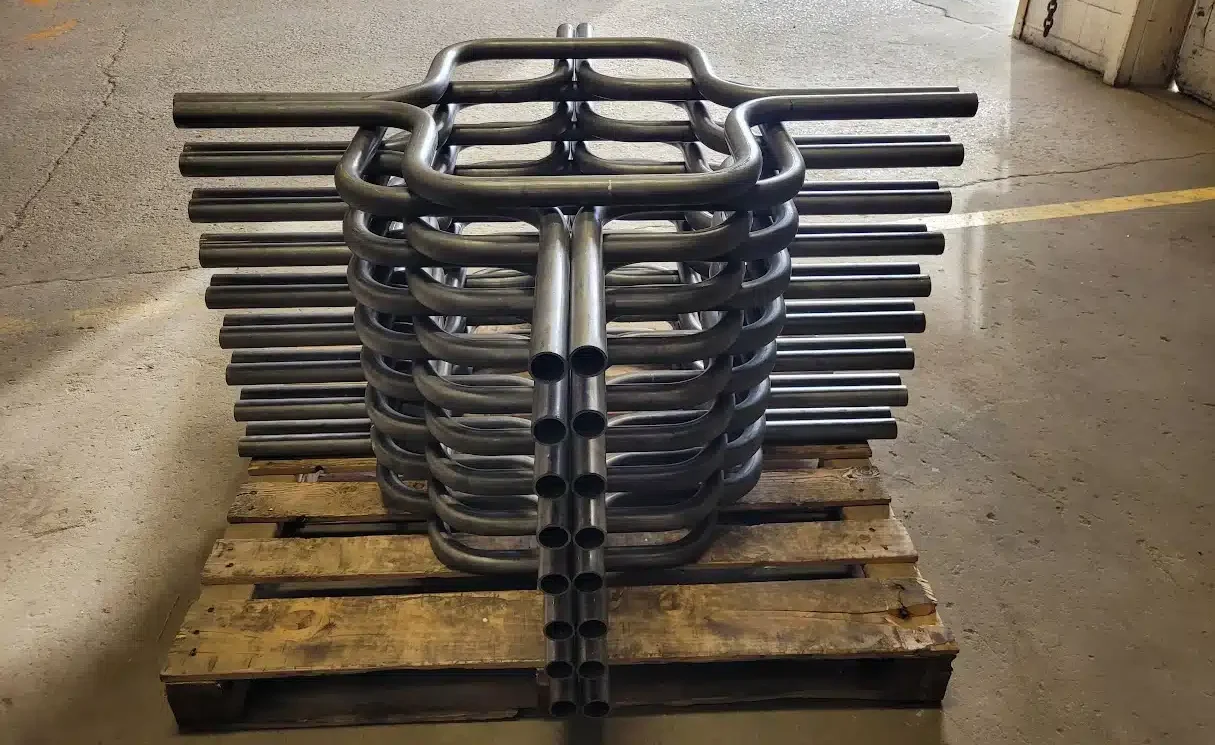 Stacked metal pipes with curved ends, produced using CNC Tube Bending, are arranged neatly on a wooden pallet in a warehouse, with an open garage door and part of a car visible outside.