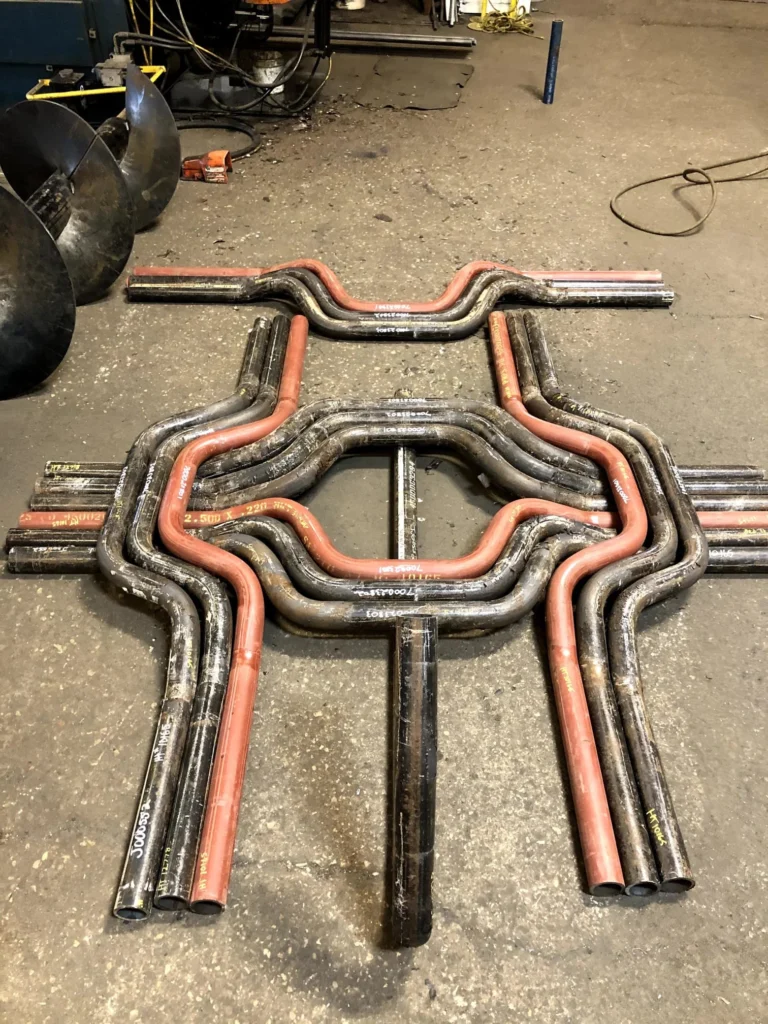 Several curved and straight metal pipes, some black and some red, are arranged on a concrete floor in a symmetrical, geometric pattern, showcasing the precision of CNC tube bending in creating striking mechanical structures or abstract sculptures.