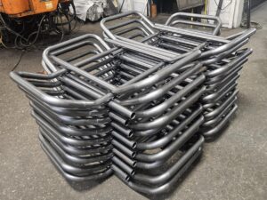 A stack of identical bent metal pipes, the result of precision tube bending, is neatly arranged on a workshop floor in Calgary, with industrial equipment and various tools visible in the background.