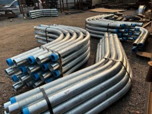 Bundles of large radius conduit, curved metal pipes with blue plastic caps are stacked on a gravel yard, surrounded by other metal materials and vehicles in the background.