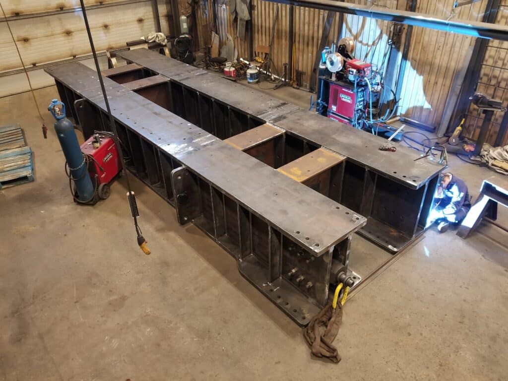 A person welds a large metal structure in a workshop, surrounded by welding equipment, tools, and a gas cylinder on a concrete floor—preparing essential components often used in Raisebore Drilling operations.