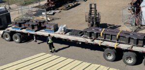 A worker in a high-visibility vest stands near a flatbed lorry loaded with large metal parts used for raisebore drilling in an industrial yard, surrounded by fencing, stacked materials, and a forklift truck.