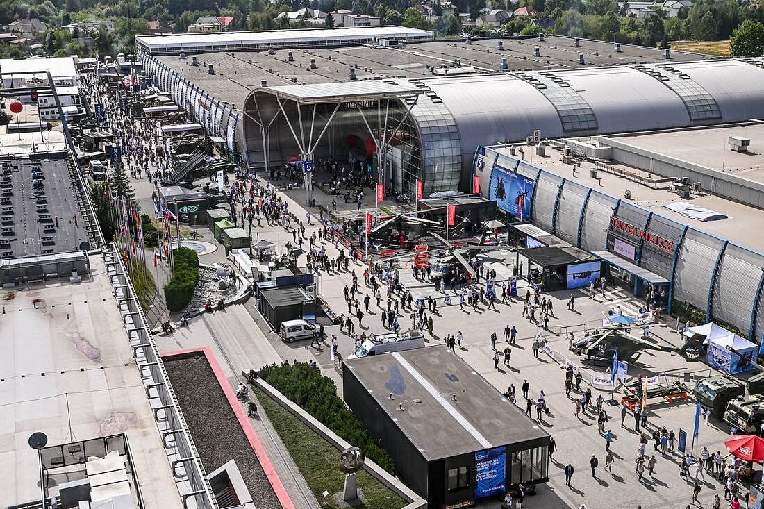 Aerial view of the bustling MSPO 2026 exhibition centre, with crowds walking among large halls, outdoor displays, and various stands. Some military vehicles are visible, and trees surround the venue.