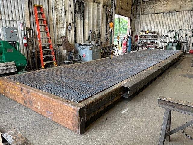 Large metal platform with grated steel surface designed to support Raisebore Drilling operations inside an industrial workshop, surrounded by tools, machinery, a red ladder, and corrugated metal walls.