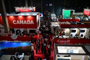Crowd walking through a busy trade fair or conference with multiple red and white Canada stands, company displays, and banners in a large indoor venue.