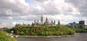 A panoramic view of Parliament Hill in Ottawa, Canada, with historic buildings crafted by skilled Canadian fabricators, surrounded by trees and set against a cloudy sky and the Ottawa River in the foreground.