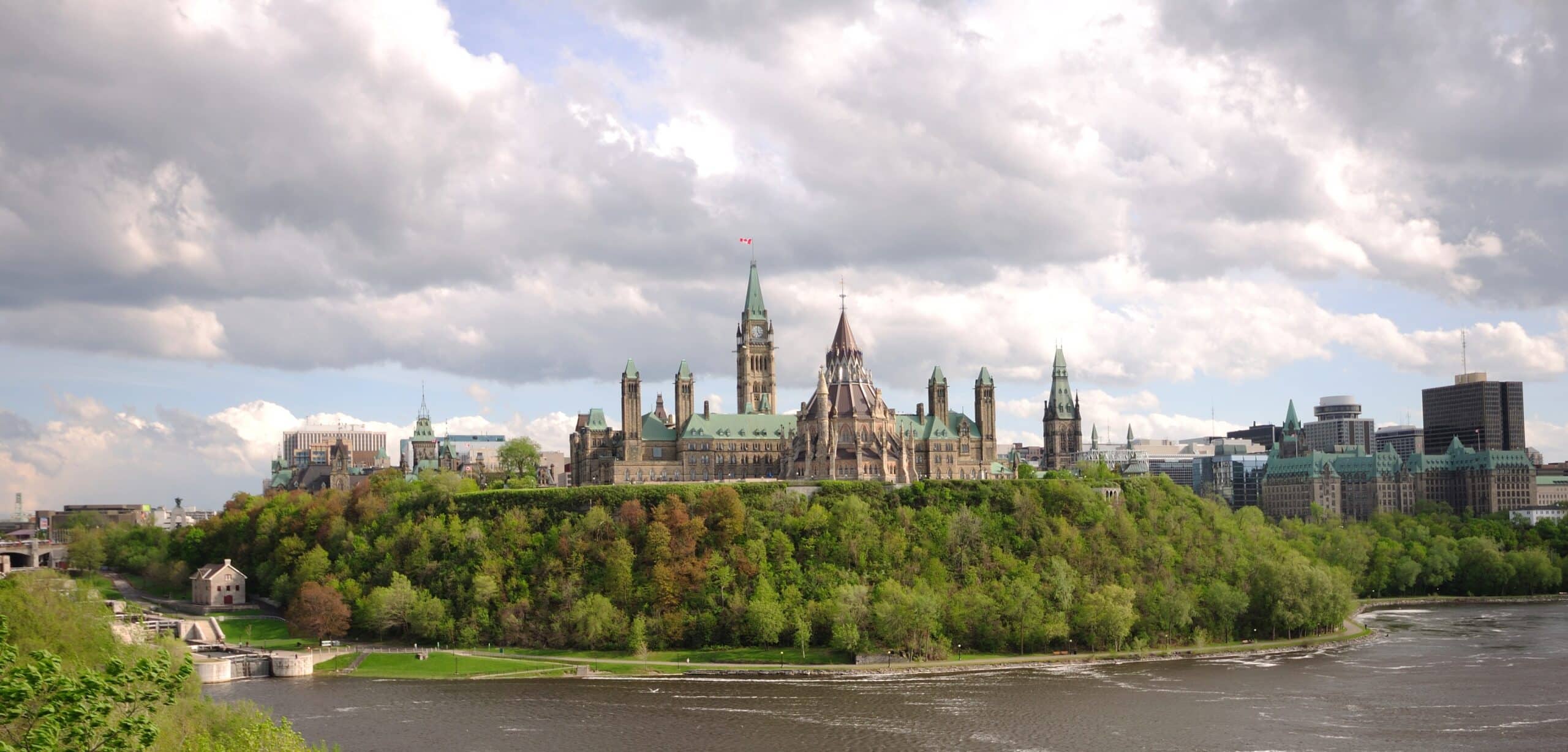 A panoramic view of Parliament Hill in Ottawa, Canada, with historic buildings crafted by skilled Canadian fabricators, surrounded by trees and set against a cloudy sky and the Ottawa River in the foreground.