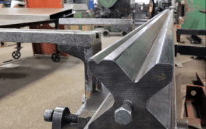 Close-up of a metal workpiece in a workshop, clamped to a metal table. The workpiece has a shiny, machined surface with a V-shaped groove, surrounded by industrial equipment and tools—a striking example of precision manufacturing amid ongoing discussions about steel tariffs.
