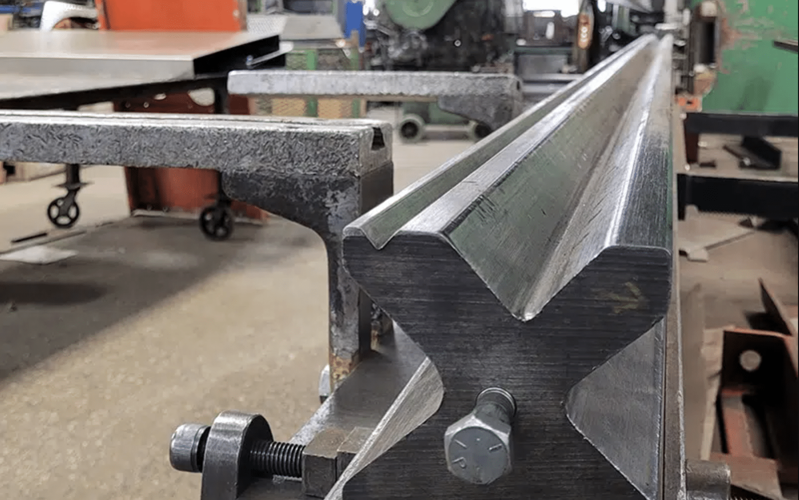 Close-up of a metal workpiece in a workshop, clamped to a metal table. The workpiece has a shiny, machined surface with a V-shaped groove, surrounded by industrial equipment and tools—a striking example of precision manufacturing amid ongoing discussions about steel tariffs.